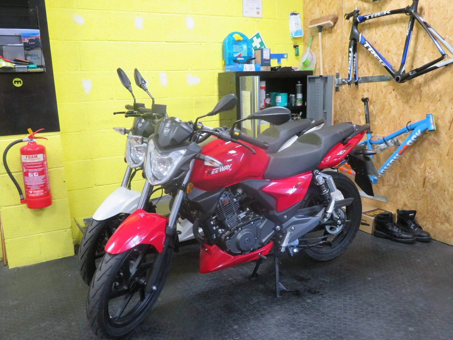 A red motorbike on the workshop floor at Veloheads, with bikes and tools on the bright yellow walls behind it